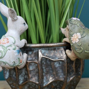 May include: A white rabbit figurine with floral details and a green hedgehog figurine with floral details are perched on the edge of a brown metal planter. Both figurines have a whimsical, garden-themed design.