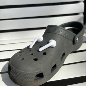 Puede incluir: Un par de auriculares inalámbricos blancos están metidos en los agujeros de una zapatilla de color gris oscuro.