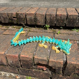 Könnte beinhalten: Ein türkis-gelbes, bewegliches Drachenspielzeug liegt auf einer Ziegeloberfläche. Der Drache hat ein detailliertes Design mit einem stacheligen Rücken und einem detaillierten Kopf. Das Spielzeug ist aus Kunststoff und scheint 3D-gedruckt zu sein.