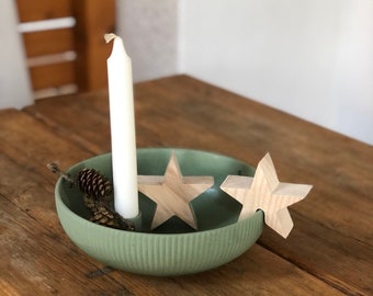 May include: A green ceramic bowl with a white candle and two wooden star ornaments. The bowl is on a wooden table.