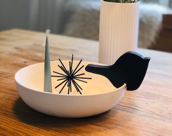 May include: A white ceramic bowl with a black wooden bird-shaped accent and a black spiky decorative object. A tall, light green candle and a white vase with branches and greenery are also present. The objects are on a wooden table.