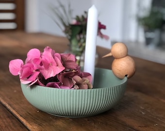 May include: A green ceramic bowl with a ribbed texture holds pink hydrangeas and a white candle. A wooden bird figurine with a bell sits on the rim of the bowl.