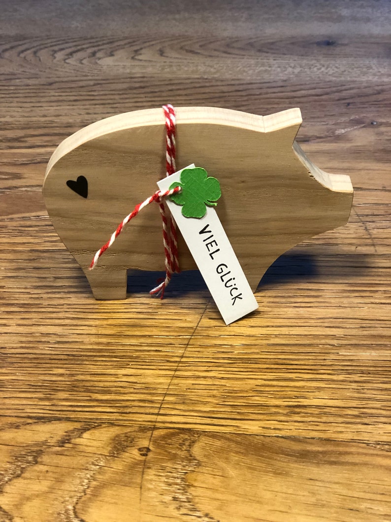 May include: Wooden piggy bank with a red and white string tied around it. A green shamrock and a tag that says "VIEL GL&Uuml;CK" are attached to the string.