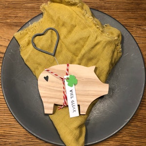May include: A wooden pig figurine with a red and white string tied around it. The pig is holding a small white tag with the text "VIEL GL&Uuml;CK" written on it. The pig is sitting on a gray plate with a yellow cloth napkin.