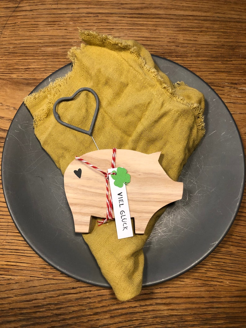 May include: A wooden pig figurine with a red and white string tied around it. The pig is holding a small white tag with the text "VIEL GL&Uuml;CK" written on it. The pig is sitting on a gray plate with a yellow cloth napkin.