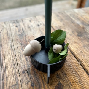 May include: A black candle holder with a single teal candle and two wooden acorn decorations. Green leaves are arranged around the candle and acorns.