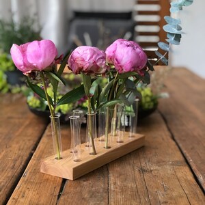 May include: A wooden flower vase with 8 clear glass test tubes for holding stems. The vase is filled with pink peonies and green eucalyptus.