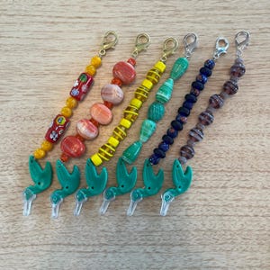 May include: Six colorful beaded eyeglass chains with gold-tone clasps. Each chain features a unique combination of glass beads in various shapes and colors, including red, orange, yellow, green, blue, and purple. The chains have a green hummingbird charm.