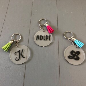 May include: Three clear acrylic keychains with black lettering and colorful tassels. The keychains feature the letters "K", "L", and the text "NDLPT".