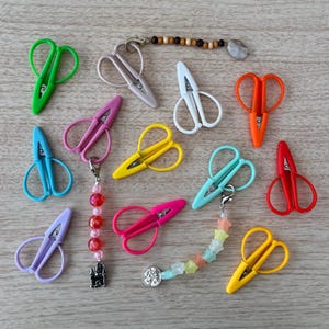 May include: A collection of small, colorful craft scissors in various shades, including green, pink, yellow, and blue. Some scissors have decorative charms attached, such as beads and a castle pendant. The scissors are arranged on a light wooden surface.