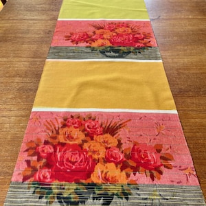 May include: A rectangular table runner with a floral design. The runner features alternating bands of yellow, pink, and floral patterns with red and orange roses. The floral pattern is set against a black and white striped background.