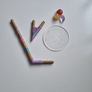 Può includere: Due bastoncini di legno con filo viola, rosa e blu avvolto attorno. I bastoncini sono piegati a forma di V. Nell'angolo in alto a destra dell'immagine ci sono tre piccole pietre: una arancione, una gialla e una viola. Un adesivo rotondo trasparente con il testo "Self Ceremony" e un simbolo si trova nell'angolo in basso a destra dell'immagine.