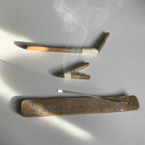 Könnte beinhalten: Ein goldfarbener Metall-Räucherofen mit floralem Design und einem grauen Räucherstäbchen. Zwei Holz-Räucherstäbchen sind ebenfalls zu sehen, eines mit einem weißen und grünen Streifenmuster und das andere mit einem weißen und braunen Streifenmuster. Rauch steigt aus dem Räucherstäbchen auf.