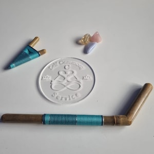 Può includere: Un timbro di vetro trasparente con il testo "Self Compassion Service" e un simbolo di infinito stilizzato con una "C" al centro. Due bastoncini di legno con una corda blu e verde avvolta intorno sono su una superficie bianca. Tre piccoli cristalli si trovano nell'angolo in alto a destra dell'immagine.
