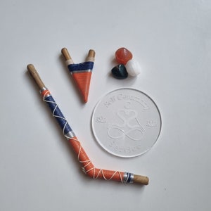 Può includere: Un assortimento di oggetti spirituali tra cui una bacchetta di legno avvolta, una piccola fionda, un sottobicchiere circolare bianco con la scritta "Self Ceremony Service" e una collezione di pietre levigate.