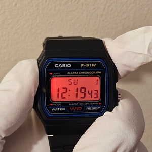 May include: A black Casio F-91W digital watch with a red digital display showing the time 12:1943 and the day of the week SU. The watch has a blue border around the display and the words 'CASIO', 'F-91W', 'ALARM CHRONOGRAPH', 'LIGHT', 'MODE', 'WATER', 'WR', 'RESIST', and 'ALARM ON-OFF/24HR' printed on the face.