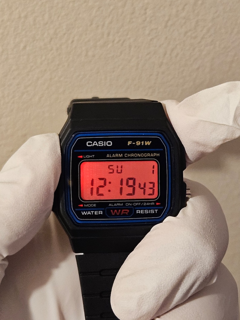 May include: A black Casio F-91W digital watch with a red digital display showing the time 12:1943 and the day of the week SU. The watch has a blue border around the display and the words 'CASIO', 'F-91W', 'ALARM CHRONOGRAPH', 'LIGHT', 'MODE', 'WATER', 'WR', 'RESIST', and 'ALARM ON-OFF/24HR' printed on the face.