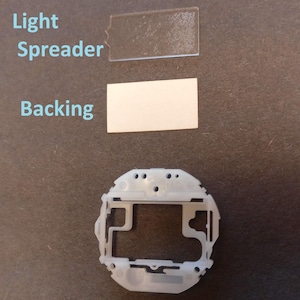 May include: An overhead shot of disassembled electronic components. The image includes a clear, rectangular light spreader, a white backing, and a white plastic frame. The components are arranged on a dark surface, with the text "Light Spreader" and "Backing" visible.