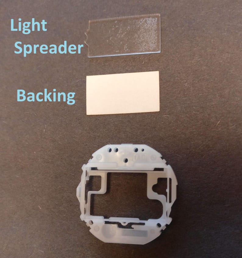 May include: An overhead shot of disassembled electronic components. The image includes a clear, rectangular light spreader, a white backing, and a white plastic frame. The components are arranged on a dark surface, with the text "Light Spreader" and "Backing" visible.