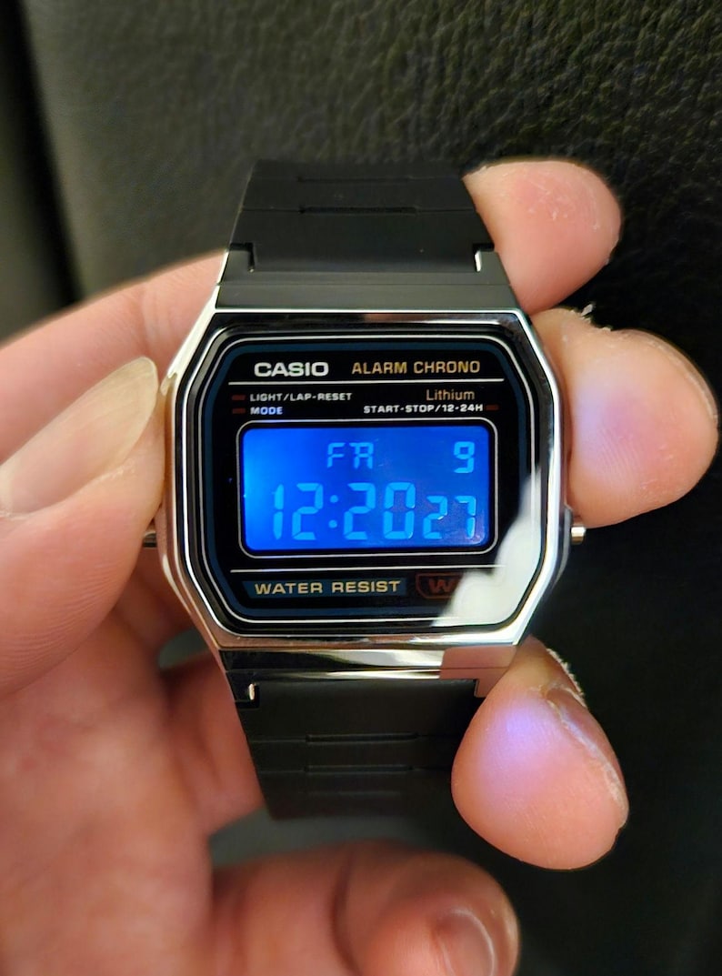 May include: A black digital wristwatch with a silver trim. The watch face displays the time 12:2027, the day of the week FR, and the number 9. The watch also has the words 'CASIO ALARM CHRONO', 'LIGHT/LAP-RESET', 'Lithium', 'MODE', 'START-STOP/12-24', 'WATER RESIST W' printed on the face.