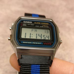 May include: A silver Casio digital watch with a black and blue strap. The watch face displays the time 11:14:54, the day, and the date. The watch has a blue and black bezel with the words "WATER RESIST" and "WR".