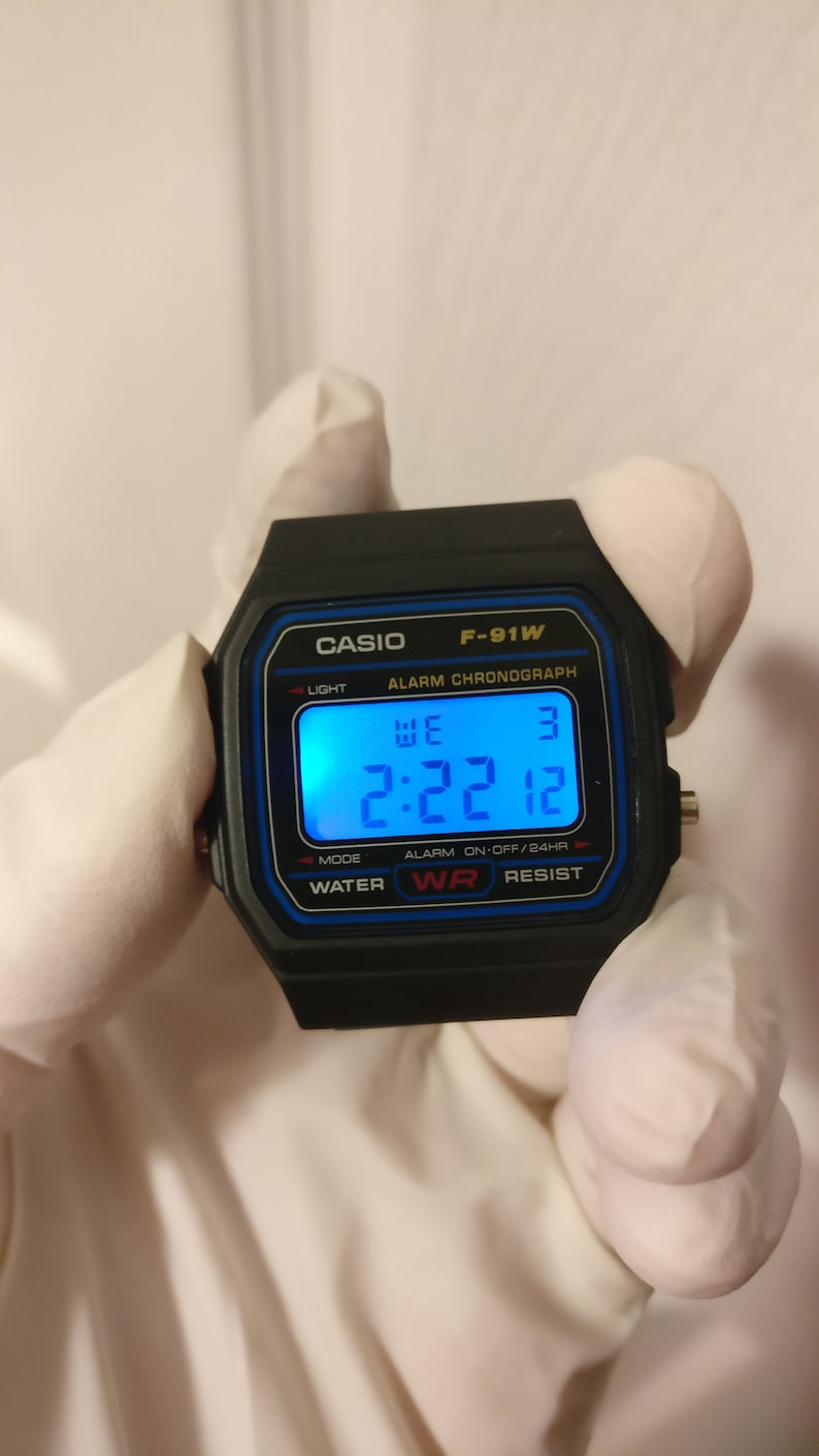 May include: A black Casio F-91W digital watch with a blue display. The watch face shows the time 2:22, the day of the week 'WE', and the date '3'. The watch has a water resistant feature and an alarm chronograph.