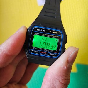 May include: A black Casio F-91W digital watch with a green display showing the time 1:08:34. The watch has a blue and black bezel and a black band. The watch is water resistant and has an alarm function.