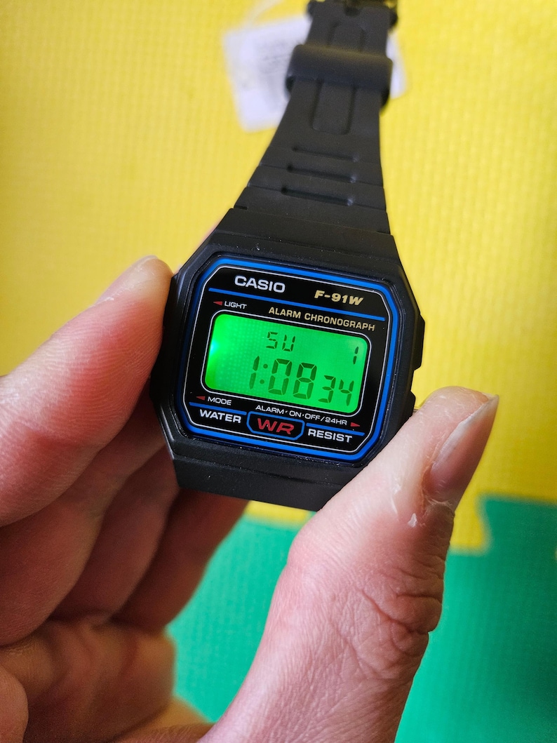 May include: A black Casio F-91W digital watch with a green display showing the time 1:08:34. The watch has a blue and black bezel and a black band. The watch is water resistant and has an alarm function.
