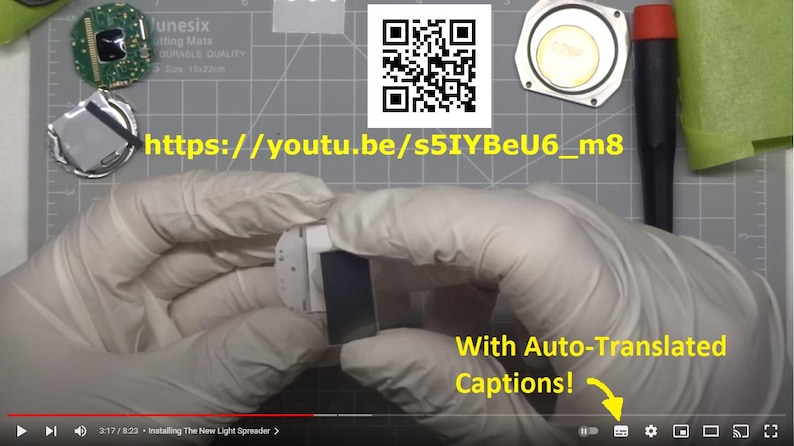 May include: A person wearing white gloves is holding a small, white electronic component with a black screen. Other disassembled electronic parts and a QR code are visible. Text on the screen reads "With Auto-Translated Captions!" and a YouTube URL is visible.