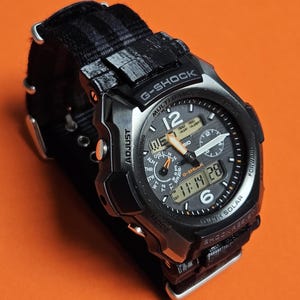 Könnte beinhalten: Eine schwarze G-Shock-Uhr mit digitaler Anzeige der Uhrzeit. Die Uhr hat ein schwarzes Armband und eine silberne Lünette. Das Zifferblatt zeigt den Text "G-SHOCK" und "MULTI BAND 6". Die Uhr ist stoßfest.