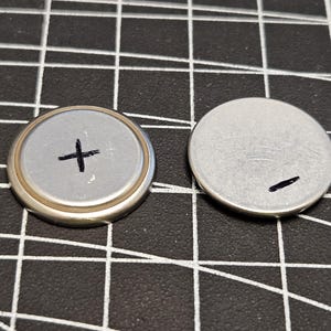 May include: Two silver-colored button cell batteries. One battery has a plus sign drawn on it with a black marker. The other battery has a minus sign drawn on it with a black marker. The batteries are on a black surface with a white grid pattern.