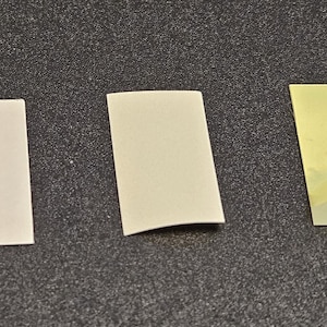 May include: Three small, rectangular adhesive pads. Two are off-white, and one has a yellow backing with a clear adhesive layer partially peeled back. The pads are arranged on a dark gray surface.