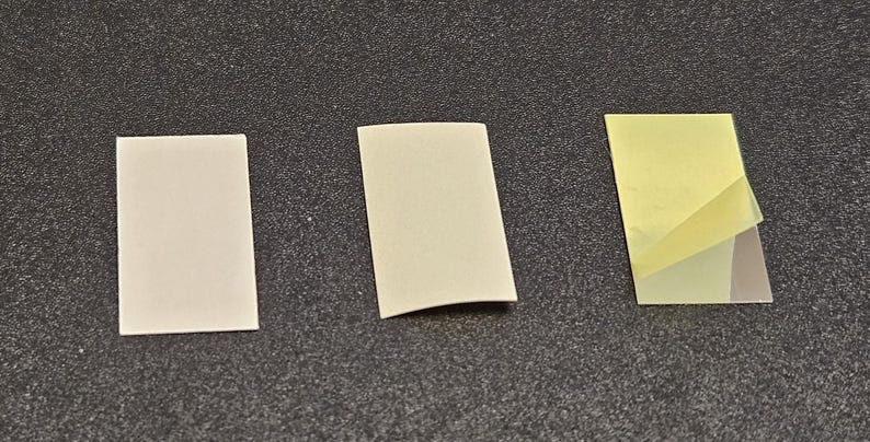 May include: Three small, rectangular adhesive pads. Two are off-white, and one has a yellow backing with a clear adhesive layer partially peeled back. The pads are arranged on a dark gray surface.