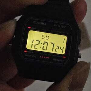 May include: A black Casio F-91W digital watch with a yellow digital display showing the time 12:07:24 and the day of the week 'SU'.