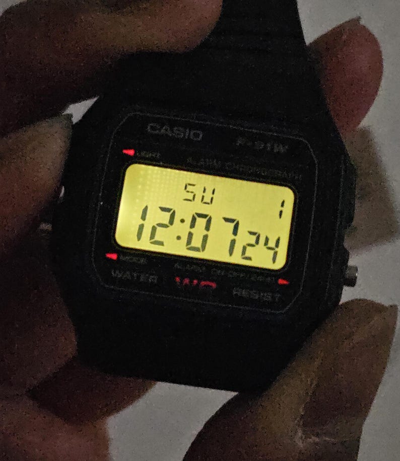 May include: A black Casio F-91W digital watch with a yellow digital display showing the time 12:07:24 and the day of the week 'SU'.