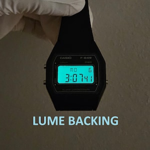 May include: A black Casio F-94W digital watch with a glowing green display showing the time. The watch face displays the day and date. The text "LUME BACKING" is below the watch.