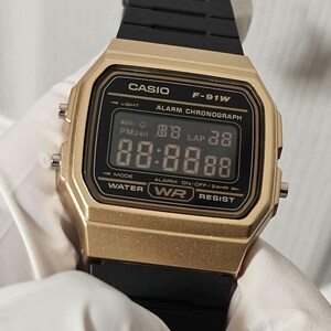 May include: A gold-colored Casio F-91W digital watch with a black band. The watch face displays the time, date, and alarm settings. The watch is water resistant.