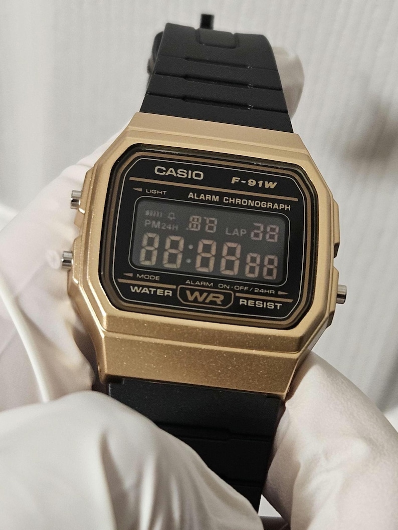 May include: A gold-colored Casio F-91W digital watch with a black band. The watch face displays the time, date, and alarm settings. The watch is water resistant.