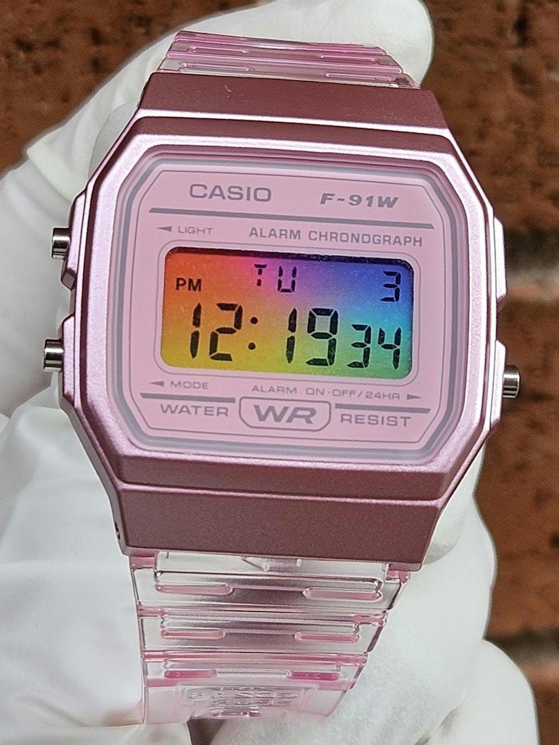May include: A pink Casio F-91W digital watch with a clear plastic band. The watch face has a rainbow background and displays the time, date, and day of the week. The watch also has an alarm and chronograph function.