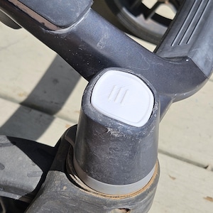 以下が含まれることがあります： ベビーカーフレームの一部のクローズアップ。フレームは濃いグレーで、3本の縦線が入った白いボタンが付いています。車輪やベビーカーの他の部分が背景に見えます。