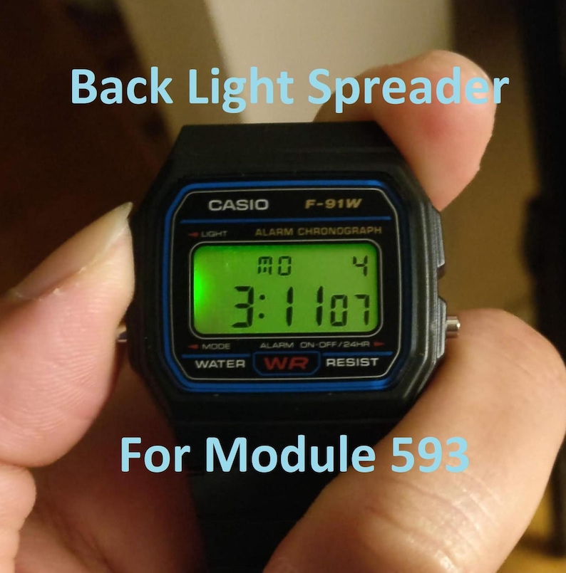 May include: Black Casio F-91W digital watch with a green illuminated display showing the time. The watch face displays "CASIO F-91W" and "Back Light Spreader For Module 593". The watch is held in a hand.