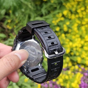 May include: Black Casio G-Shock watch with a black strap and silver buckle. The watch face is visible, with the words "Shock Resistant" and "Casio" printed on it. The watch is held against a background of green foliage and yellow flowers.