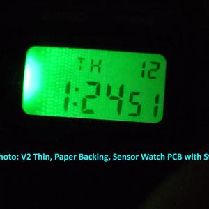 May include: A close-up of a digital display with a green backlight. The display shows the time as 1:24:51, with the day and date above. The text "In this photo: V2 Thin, Paper Backing, Sensor Watch PCB with Stock LCD" is at the bottom.