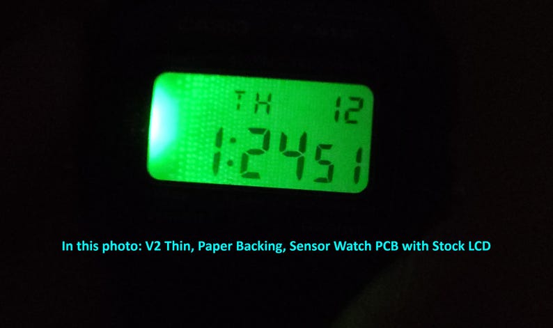 May include: A close-up of a digital display with a green backlight. The display shows the time as 1:24:51, with the day and date above. The text "In this photo: V2 Thin, Paper Backing, Sensor Watch PCB with Stock LCD" is at the bottom.