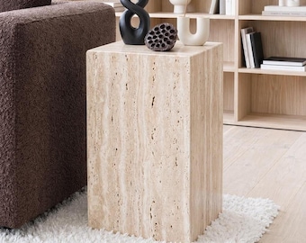 May include: A beige travertine side table with a rectangular shape. The table is placed on a white shag rug. A black sculpture, a dried lotus pod, and a white vase are on top of the table. The table is 18 inches tall.