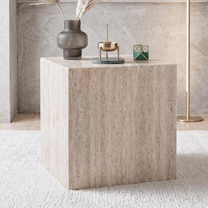 Könnte beinhalten: Ein beiger Travertin-Würfeltisch mit strukturierter Oberfläche. Eine graue Vase mit getrockneten Pflanzen, ein goldener Kerzenhalter und ein grünes geometrisches Objekt stehen darauf. Der Tisch steht auf einem weißen Teppich.