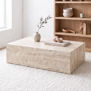 Pode incluir: Uma mesa de centro retangular de travertino bege. A mesa é um bloco sólido com uma superfície texturizada. Um pequeno vaso redondo bege com um ramo de vegetação está sobre a mesa, junto com um livro e esferas decorativas. Uma estante de madeira está ao fundo.