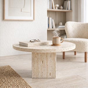 Pode incluir: Uma mesa de centro redonda de pedra bege com base quadrada. Um livro, óculos e uma caneca de café estão em cima. A mesa está numa sala de estar com uma cadeira e uma estante.