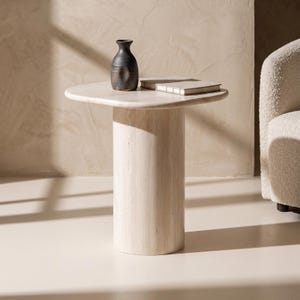 May include: A beige travertine side table with a cylindrical base and a rounded square top. A dark charcoal ceramic vase and a closed rectangular book with a white cover are placed on the surface of the table, which has a smooth, porous texture.