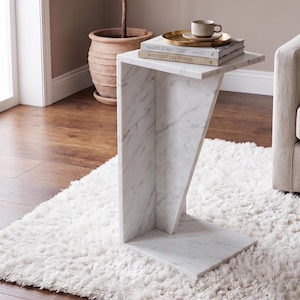 Op de afbeelding: Een C-vormige bijzettafel van wit marmer met grijze aders. De tafel heeft een plat bovenoppervlak, een verticaal steunpaneel en een platte voet. Twee boeken met de titels Marble Mastery en Mid-Century Forms liggen bovenop met een gouden dienblad en een kopje.